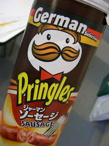 Three Pringles Pop Dog Buns in their iconic cans, with flavor labels for BBQ, Sour Cream & Onion, and Honey Mustard. A hot dog in one of the buns is visible.